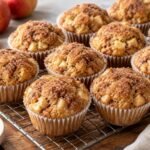 Cinnamon Apple Muffins: A batch of golden cinnamon apple muffins in white paper liners on a cooling rack, domed tops coated with sparkling cinnamon sugar, small pieces of apple visible at the edges.