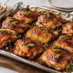 Honey Garlic Chicken Thighs: Glossy honey garlic chicken thighs on a foil-lined sheet pan straight from the oven, sticky caramelized glaze with browned edges, sliced green onion scattered on top.